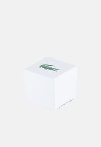 Valge ruudukujuline kast tekstureeritud pinnaga, mille peal on roheline krokodilli logo ja külgedel on trükitud "LACOSTE".