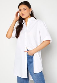 Camecia bianca a maniche corte con bottoni, realizzata in morbido tessuto di cotone, con orlo dritto e spacco laterale. Abbinata a jeans blu.