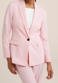 Helles rosa, maßgeschneidertes Blazer mit einem einzelnen Knopfverschluss, mit Schößchen und zwei vorderen Taschen. Getragen über einem weißen Oberteil und passenden Hosen.