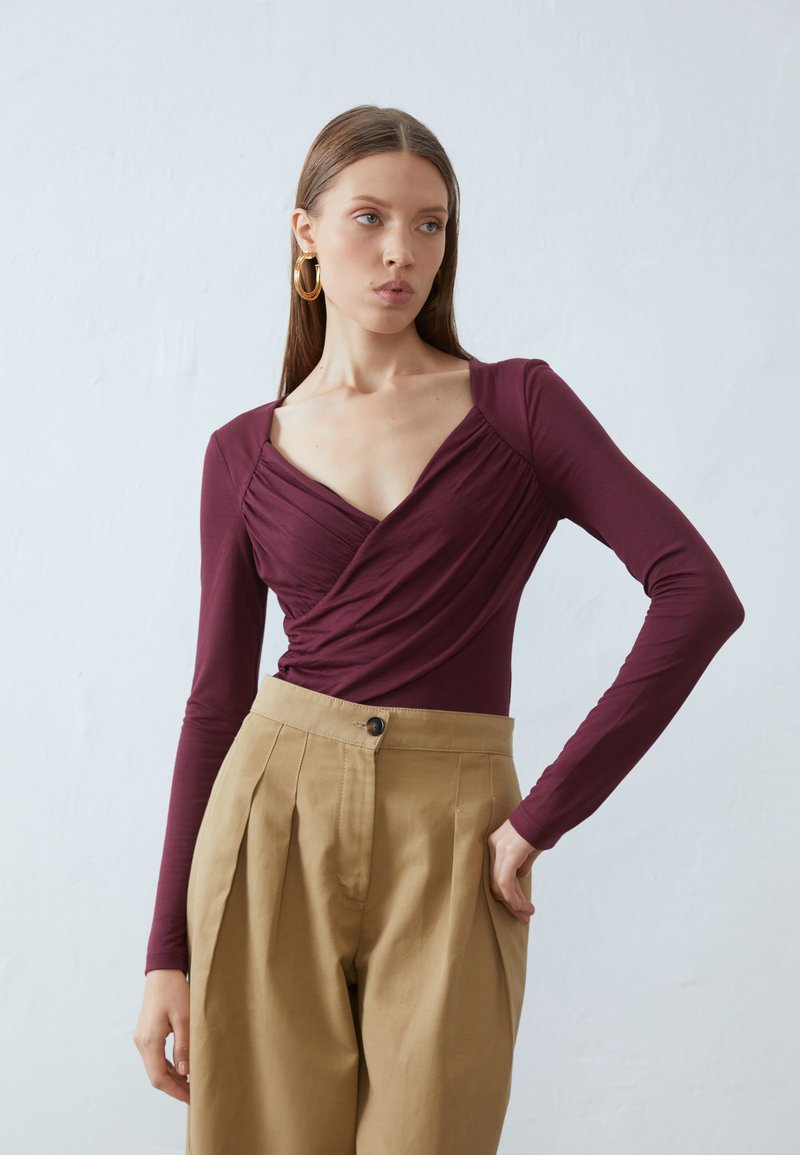 Burgundy long-sleeve wrap top with ruched detailing, paired with tan high-waisted pleated trousers. Smooth texture and fitted design.