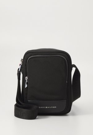 Musta kompakti olkalaukku edessä olevan vetoketjutaskun, säädettävän olkahihnan ja Tommy Hilfiger -logon kanssa etupaneelissa.