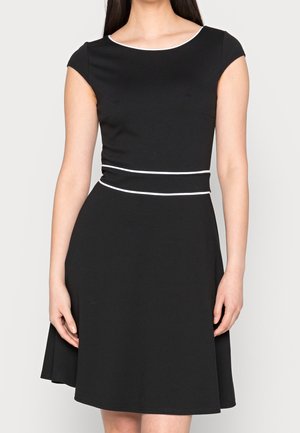 Kobieta w czarnej sukience o długości do kolan z krótkimi rękawami typu „capa” i białym obszyciem przy dekolcie oraz talii, stojąca na jednolitym tle.