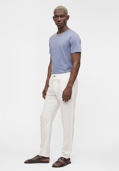 Homme mince aux cheveux blonds courts, portant une chemise bleu clair à manches courtes, un pantalon blanc à cordon et des sandales marron foncé, debout devant un fond uni.