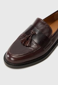 Dark brown leather loafers with a smooth finish, featuring a tassel detail and a contrasting leather band across the upper. Rubber sole.
