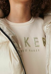 Personne portant une veste matelassée beige sur un sweat crème avec le texte métallique « BAKER TED BAKER ». Cheveux bouclés partiellement visibles.