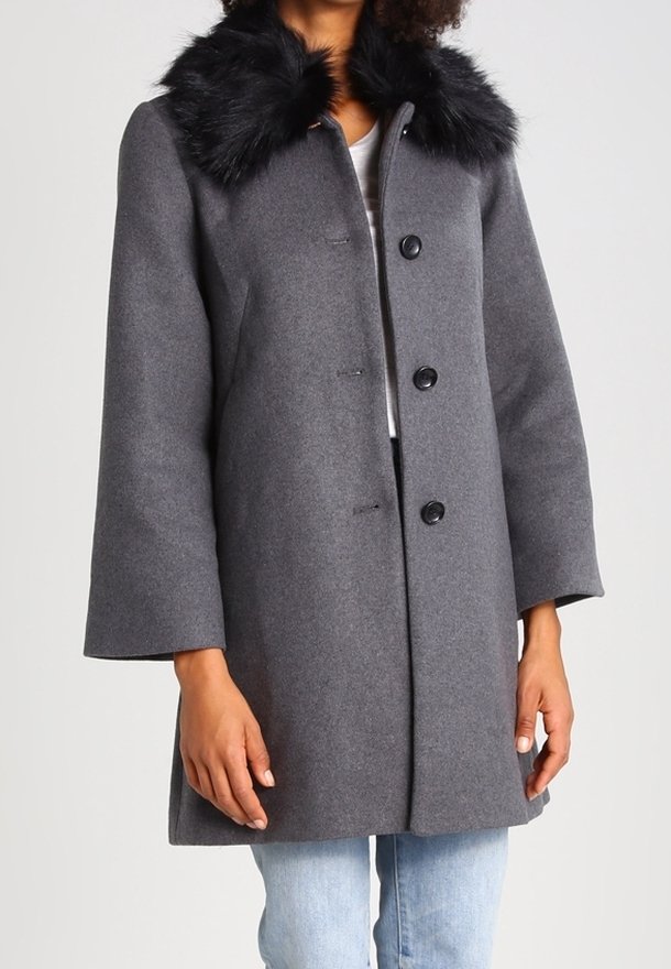 Femme portant un manteau gris mi-long avec un col en fausse fourrure noire, boutonné devant et manches longues, sur un jean bleu clair.