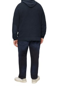 Navyblaue gestrickte Kapuzenjacke mit gerippter Textur, ausgestattet mit einer Kapuze und Ellbogenpatches. Kombiniert mit dunkelblauen Jeans und hellen Sneakers.