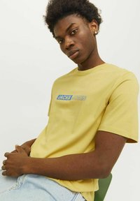 Camiseta de algodón amarilla con un logo azul y blanco de "JACK & JONES" en el pecho, cuello redondo y mangas cortas.