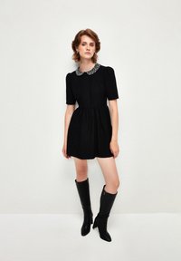 Schwarzes kurzes Kleid mit einem runden, mit Perlen verzierten Kragen, gepufften Ärmeln und einem ausgestellten Rock; kombiniert mit knielangen, schwarzen Lederstiefeln.