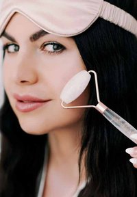 Rolletto viso in quarzo rosa con manico trasparente, utilizzato sulla pelle. Una maschera per gli occhi di colore rosa chiaro copre parzialmente i capelli scuri.