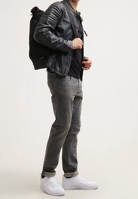 Persona vestida con una chaqueta de cuero negra, jeans gris oscuro, zapatillas blancas y llevando una mochila negra sobre un hombro, de pie de lado.