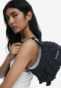 Black denim backpack with floral fabric accents, strap details, and logo. Smooth texture with layered flower embellishments.