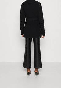 Cardigan nero a coste con cintura, dotato di cappuccio e maniche lunghe; abbinato a pantaloni a zampa lucidi neri e scarpe con tacco aperto.