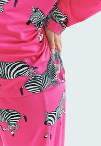 Souprava loungewear z bavlny s jasně růžovým pozadím a černobílými zebrovými vzory. Obsahuje tričko a kalhoty s viditelnou etiketou.