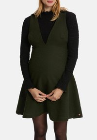Groene zwangerschapsjurk met een aansluitende corsage en uitlopende rok, gecombineerd met een zwarte textuur lange mouw top en donkere leggings.