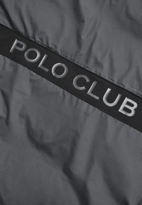 Grått tyg med en slät textur har ett framträdande svart band som visar texten "POLO CLUB" i upphöjd silverfärgad skrift.