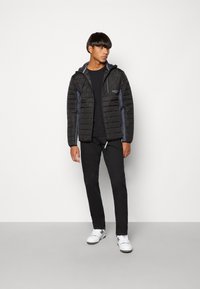 Zwarte lichtgewicht pufferjack met horizontaal gestikt ontwerp, grijze accenten, rits aan de voorkant en capuchon, gecombineerd met zwarte broek en witte sneakers.