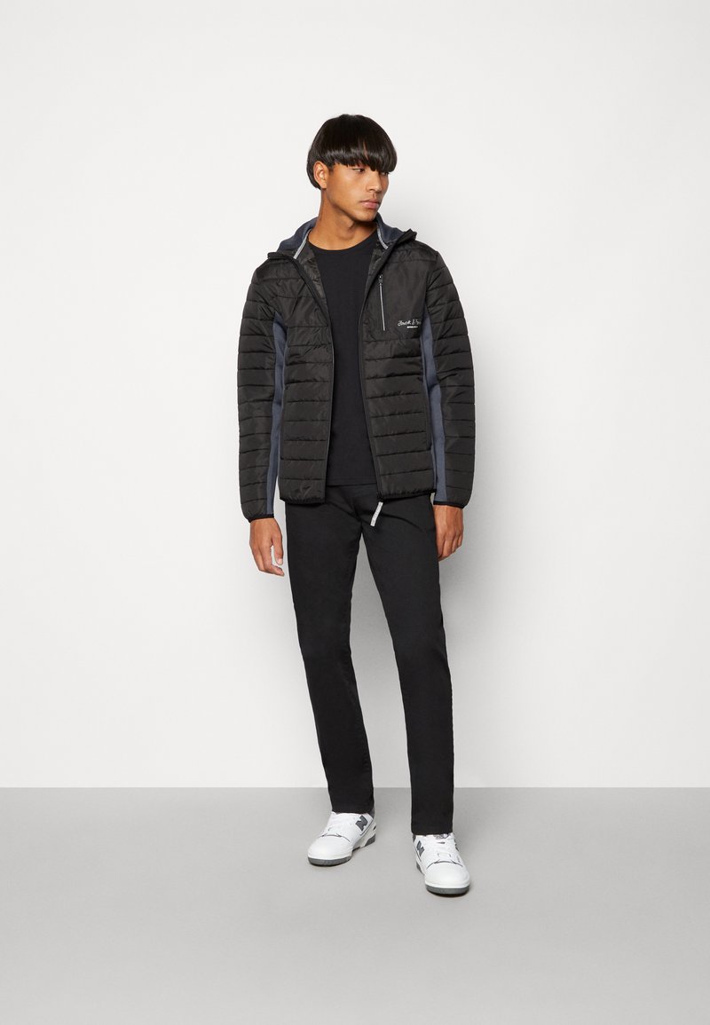 Zwarte lichtgewicht pufferjack met horizontaal gestikt ontwerp, grijze accenten, rits aan de voorkant en capuchon, gecombineerd met zwarte broek en witte sneakers.