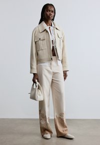 Bēša īsā jaka ar divām priekšējām kabatām, zem tā balta krekls, piemērotas bēšas platajiem šuvēm bikses ar ziedu rakstu un mazs balts rokassomiņa.