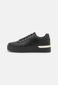 Black sneaker with textured leather upper, lace closure, and gold-tone hardware. Features a thick black sole with a metallic accent.