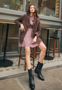 Brown suede long coat over a checkered dress with a flared skirt. Black ankle boots with studs, sitting on a wooden barstool outdoors.