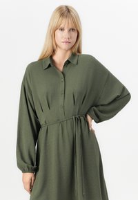 Robe vert olive en tissu texturé, avec un devant à boutons, des manches longues et une ceinture à nouer à la taille.
