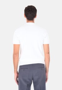 Uomo con capelli corti scuri che indossa una camicia bianca attillata a maniche corte e pantaloni grigio scuro, in piedi girato di spalle su uno sfondo neutro.