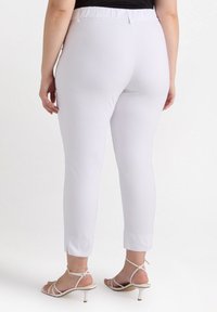 Witte, aansluitende broek met elastische tailleband, enkel lengte en een gladde textuur. Draagt met gespen sandalen op hoge hakken.