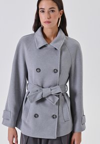 Cappotto doppio petto grigio in lana con ampio colletto, cintura, due grandi tasche frontali e bottoni decorativi. Presenta una finitura testurizzata.