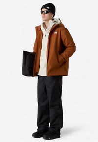 Chaqueta impermeable marrón, sudadera con capucha beige, pantalones negros, gorro negro, gafas de sol, sosteniendo una bolsa negra. Presenta el logo de The North Face en la chaqueta.