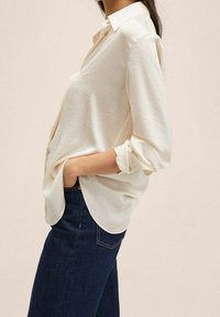 Chemise blanche à boutons, à manches longues, en tissu léger, rentrée dans un jean bleu foncé, avec manches retroussées et col classique.