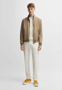 Giacca bomber marrone con zip e polsini a costine, maglione crema, pantaloni crema e sneakers gialle. Tessuto liscio con design informale.