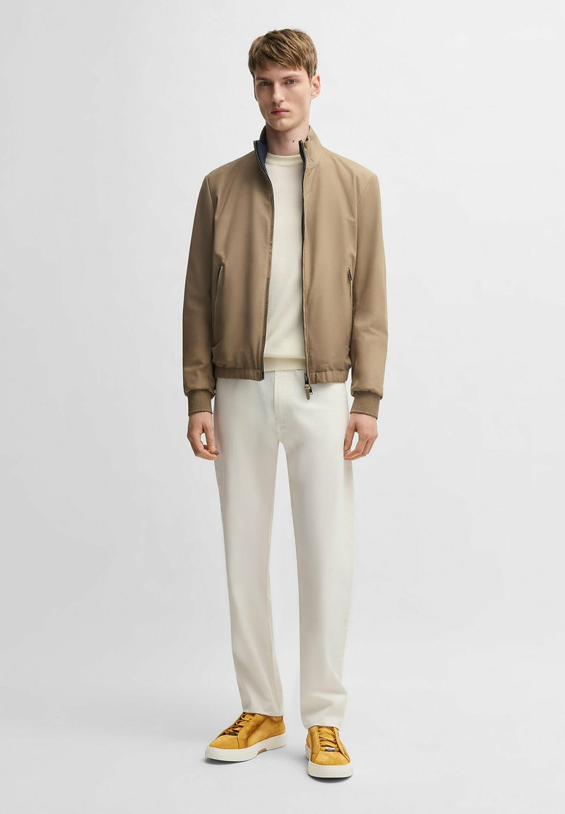 Giacca bomber marrone con zip e polsini a costine, maglione crema, pantaloni crema e sneakers gialle. Tessuto liscio con design informale.