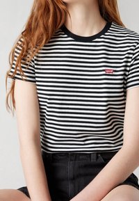 Personne aux longs cheveux roux portant un t-shirt Levi's rayé noir et blanc et un short noir taille haute, assise avec les bras reposant sur les jambes.