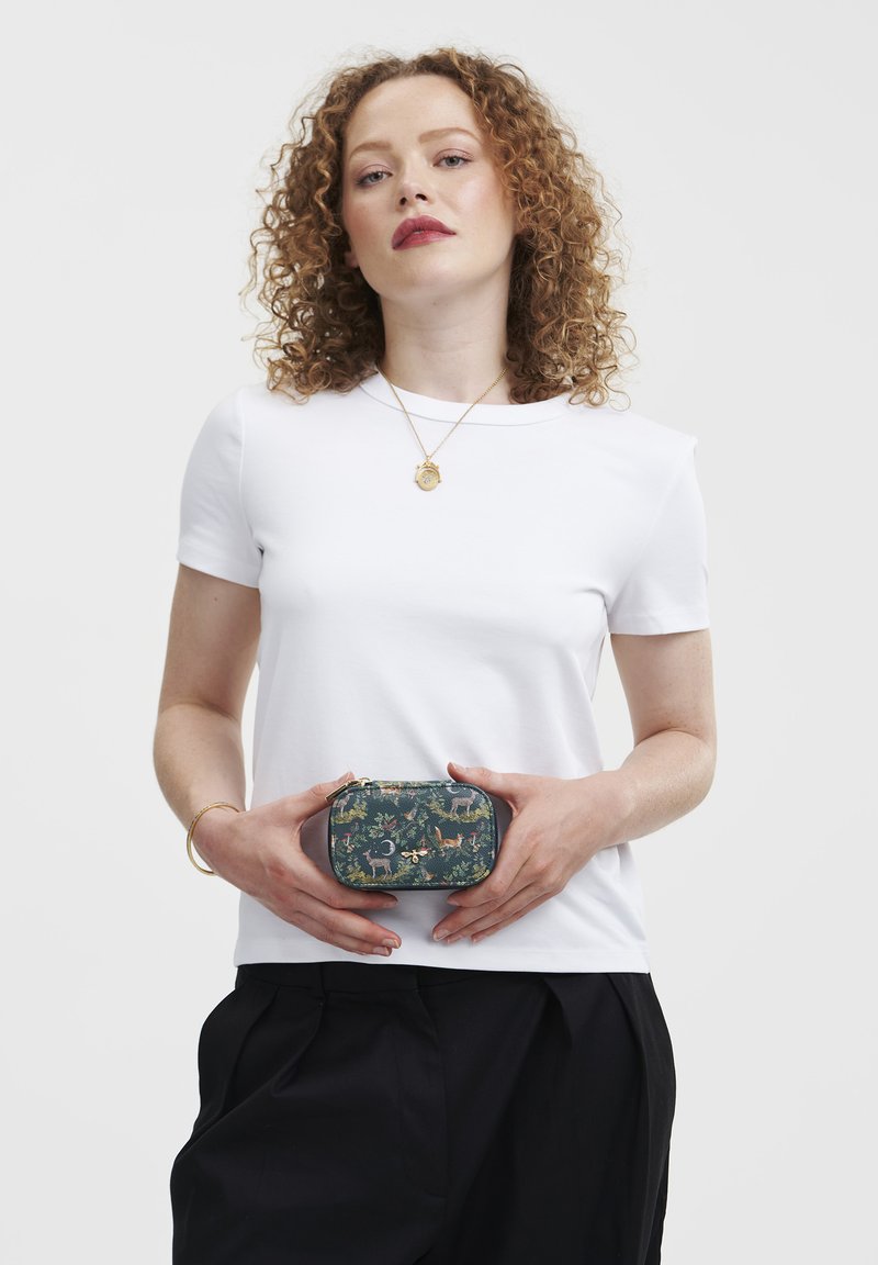 Kompakt zippered väska med ett mörkgrönt tyg som har djur- och blommönster, hålls i händerna mot en vit T-shirt och svarta byxor.