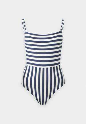 Femme debout pieds nus portant un maillot de bain une pièce avec des rayures horizontales bleu marine et blanches en haut et des rayures verticales en bas.