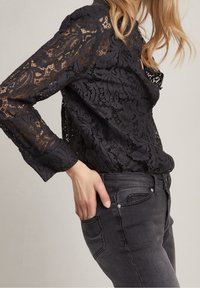 Blusa nera in pizzo con motivi floreali, maniche lunghe, polsini con bottone e un design semitrasparente, indossata con jeans grigio scuro.