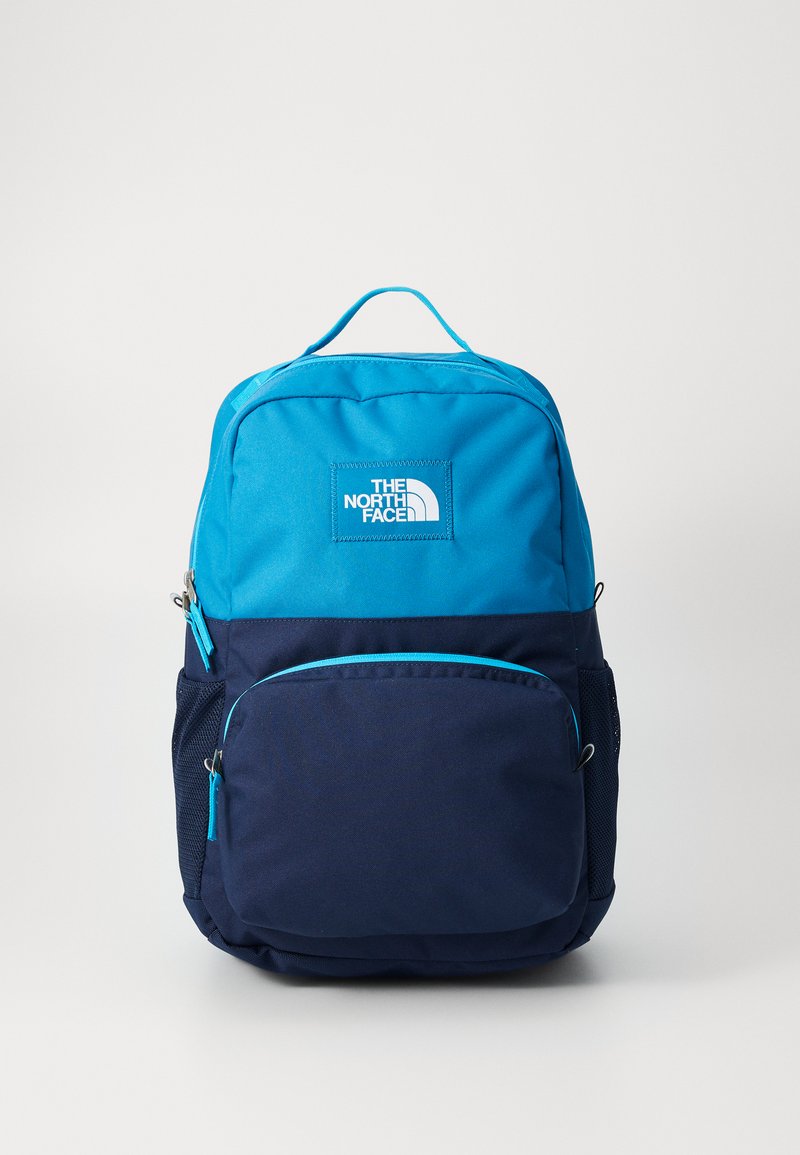 Sac à dos bleu et marine avec un design structuré, comprenant une poche zippée à l'avant, un patch logo et une poche en mesh sur le côté. Accents de fermeture éclair en bleu clair.