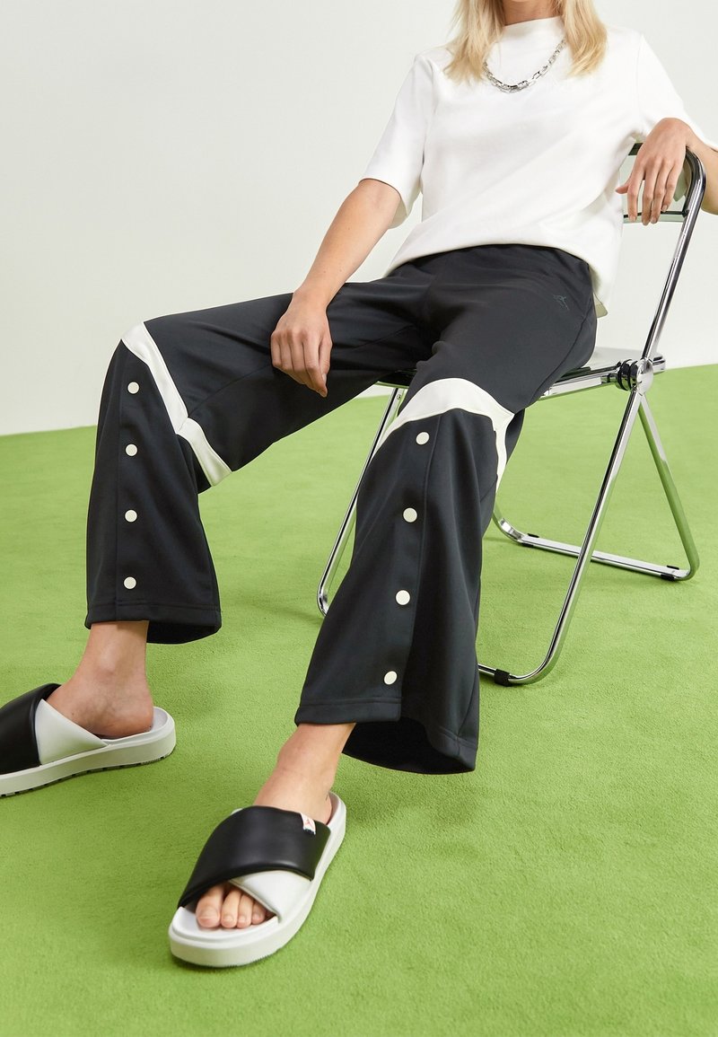 Person som bär svarta vida byxor med vita accenter och knappar, vit kortärmad topp, svarta och vita sandaler med öppen tå, sittande på en metallstol.