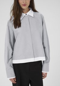 Femeie îmbrăcată cu o bluză gri lejeră, cu mâneci supradimensionate, guler și manșete albe, asortată cu pantaloni negri, pe un fundal simplu.