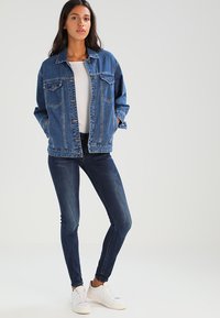 Τζάκετ τζιν σε μεσαίο μπλε πλύσιμο με χαλαρή εφαρμογή, δύο τσέπες στο στήθος και κλείσιμο με κουμπί, συνδυασμένο με σκούρες skinny τζιν και λευκά αθλητικά παπούτσια.