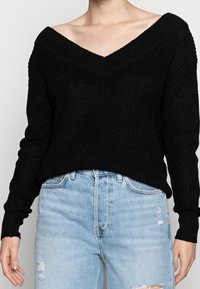 Vrouw in een zwarte off-the-shoulder gebreide trui en lichtblauwe high-waisted jeans met subtiele versleten details.