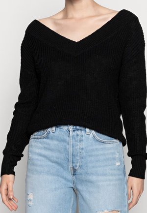 Kvinde iført en sort strikket sweater uden skuldre og lyseblå højtaljede jeans med subtile slidte detaljer.