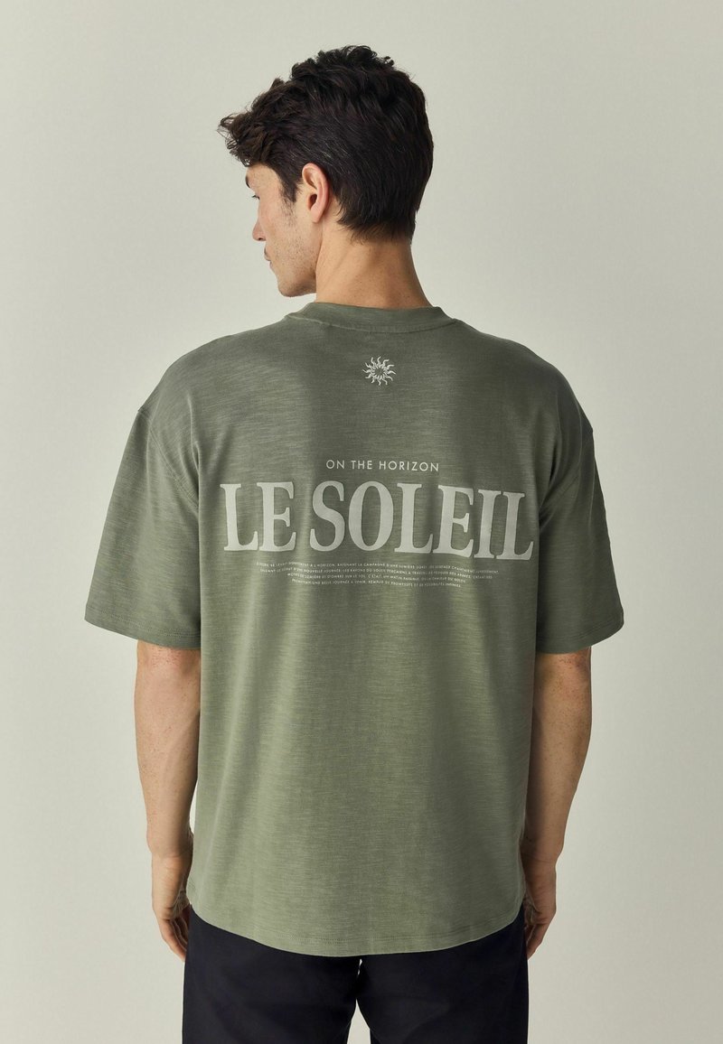 Tricou oversized de culoare verde măslin, cu textul "LE SOLEIL" și "ON THE HORIZON" imprimat în gri deschis pe spate, având un grafic cu soare deasupra.