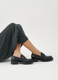 Svarte skinnloafers med kraftig såle, detaljert med en horisontal stropp over vristen. Brukt med et mørkegrønt plagg som viser jevn tekstur.