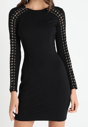 Zwarte, nauwsluitende jurk met lange mouwen, voorzien van zilveren oogjes en veterdetails langs de mouwen en schouders.