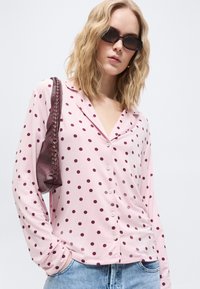 Femme portant des lunettes de soleil noires, une chemise rose à boutons avec des pois foncés, un jean bleu clair et un sac à bandoulière bordeaux, debout devant un fond uni.