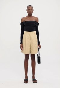 Schwarzes Off-Shoulder Langarmtop, beigetailorierte Shorts mit Taschen, schwarze Sandalen und eine kleine schwarze Handtasche. Glatte Texturen.