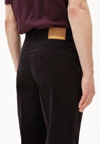 Zwarte broek met een leren label met de tekst "ARMEDANGELS" aan de achterkant van de tailleband. Gladde textuur en een nauwsluitende pasvorm met achterzakken.