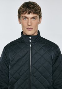 Giacca nera trapuntata con colletto alto, dotata di zip frontale e bottoni a pressione, texture liscia e dettagli a motivo diamante.